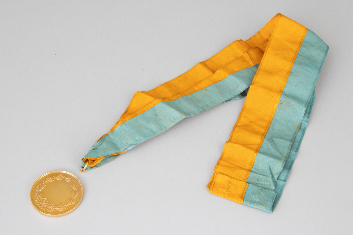 Rund medalje i forgylt metall med opphøyet rand og punslet dekor. På avers er det en globus med palmegrener rundt og en skøyte over. Innskrift langs randen. På revers er det en laurbærkrans, dannet av to kryssende grener med sløyfe rundt, ellers glatt overflate. Langt gult og turkis ripsbånd festet til en ring som er brukket av medaljens kant.