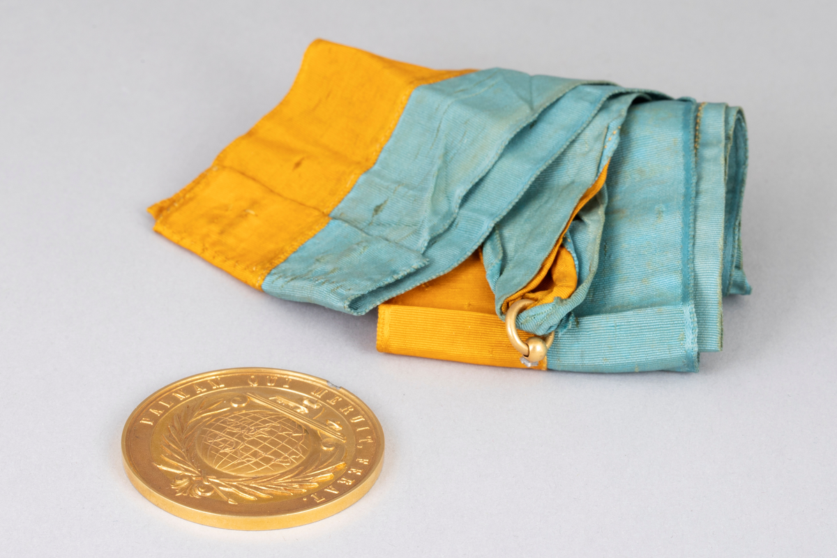 Rund medalje i forgylt metall med opphøyet rand og punslet dekor. På avers er det en globus med palmegrener rundt og en skøyte over. Innskrift langs randen. På revers er det en laurbærkrans, dannet av to kryssende grener med sløyfe rundt, ellers glatt overflate. Langt gult og turkis ripsbånd festet til en ring som er brukket av medaljens kant.