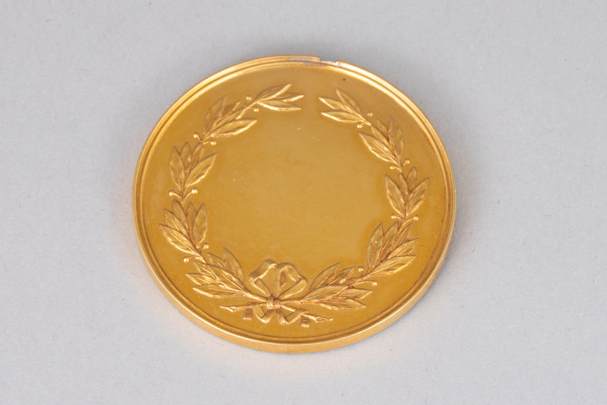 Rund medalje i forgylt metall med opphøyet rand og punslet dekor. På avers er det en globus med palmegrener rundt og en skøyte over. Innskrift langs randen. På revers er det en laurbærkrans, dannet av to kryssende grener med sløyfe rundt, ellers glatt overflate. Langt gult og turkis ripsbånd festet til en ring som er brukket av medaljens kant.