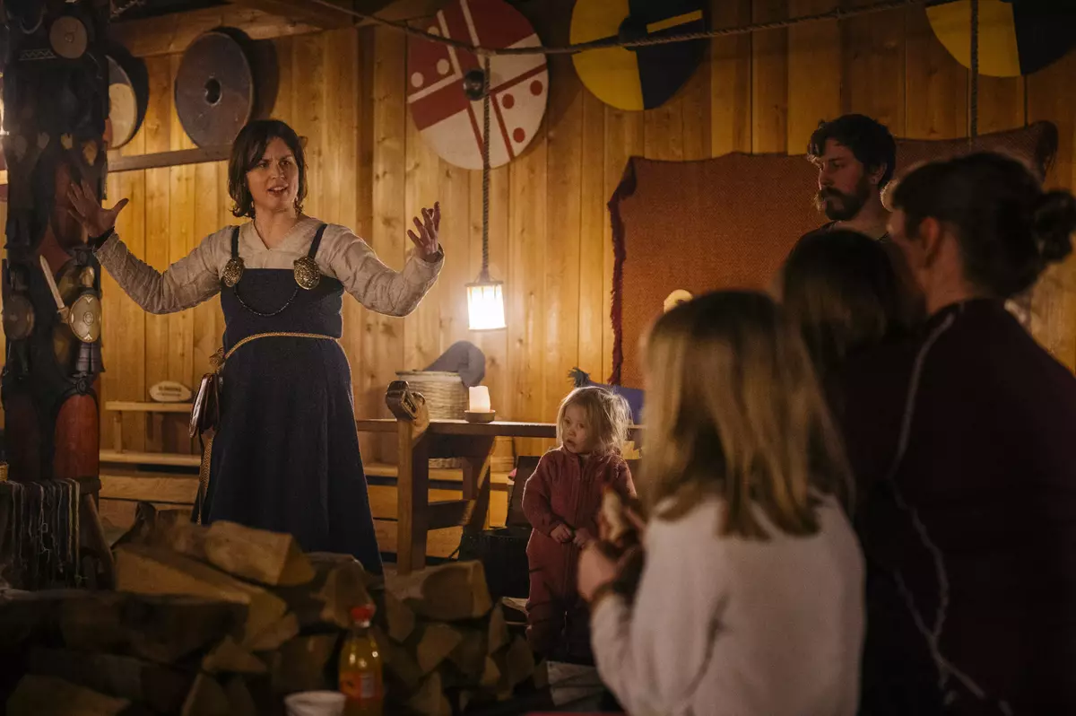 Bildet er fra Gildehallen ved Midgard vikingsenter. Det er dunkel belysninger og stemningsfullt. Til høyre sitter flere barn og følger med på en formidler som står til venstre og forteller og viser med armer. Formidleren har vikingklær på seg