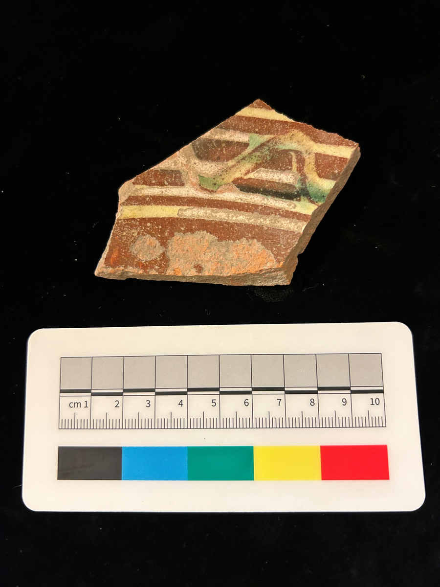 Ett fragment av yngre rödgods med brun glasyr i botten och cirklar i gult och vågband i grönt.