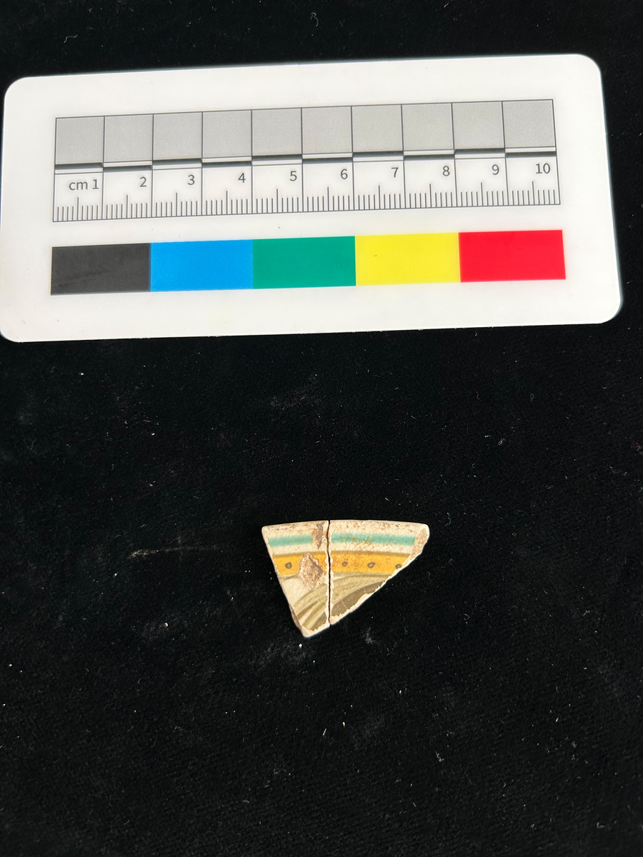 Två fragment av fat i flintgods. Bitarna har passning och är 4 mm tjocka. Glasyr i vitt, grönt, gult, orange och brunt.