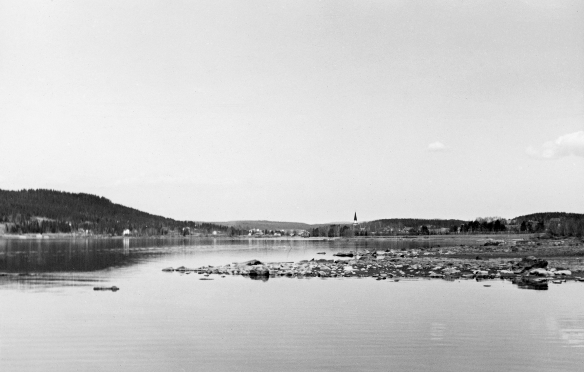 Landskapsbilde fra Sandsjøen, en sidearm av Storsjøen i Nord-Odal.  Fotografiet er tatt over ei stille, speilblank vannflate.  Et lite steinkast foran fotografen stakk et par grusskjær ut i sjøen.  I bakgrunnen ser vi et landskap med lave, skogkledde åser med litt spredt bebyggelse i strandsona ned mot sjøen.  Sentralt i bildet skimtes et kirkespir som rager mot himmelen, antakelig Sand kirke.  Ifølge en innskrift på et albumoppslag i Glomma fellesfløtingsforenings album nr. 13 skal dette og flere andre bilder på de samme albumbladene være tatt i nærheten av et bruk ved navn Svennebyhagen, som var en slags base for sjøfløtinga i Storsjøen med oppankringssted for båter og «fløtarstuggu».