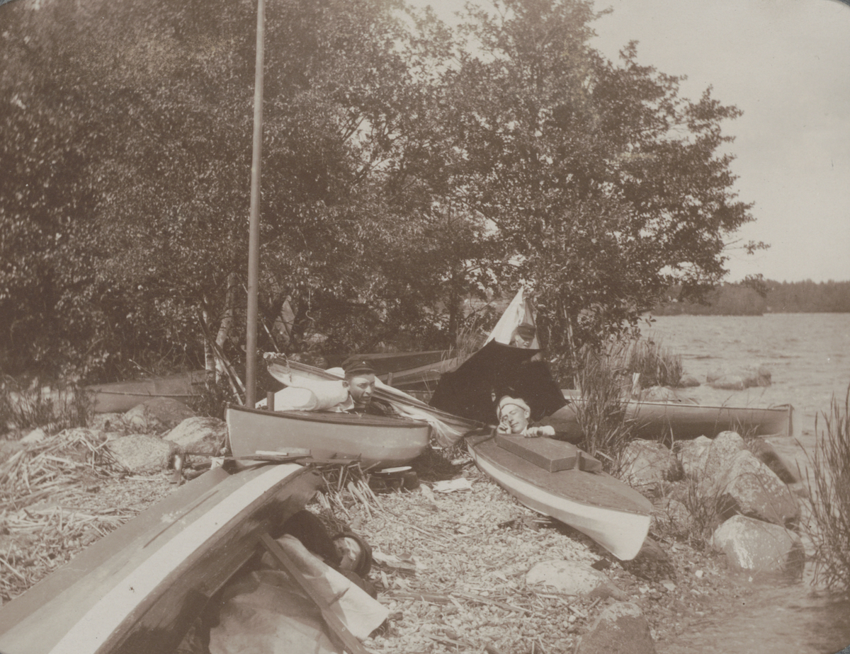 Fyra unga män vilar i kanoter som dragits upp på land. "Läger på Fredagsholmen 1906".