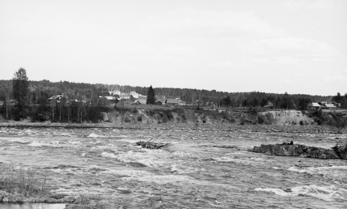 Liten tømmervase i elva, antakelig på ei av grunnene i Glomma like nedenfor kraftverksdammen ved Skjefstadfossen i Heradsbygda i Elverum.  Like nedenfor den nevnte haugen, som befinner seg noenlunde midt i bildeflata, lå det et lite steinskjær.  Over elvebrinken på motsatt side skimter vi ei grend med flere små bruk, omgitt av barskog. 