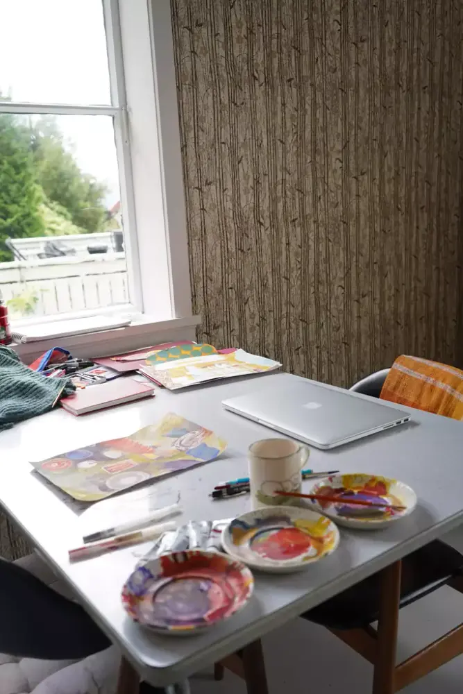 Interiør med bord og vindu i skolehuset. Malesaker på bordet.