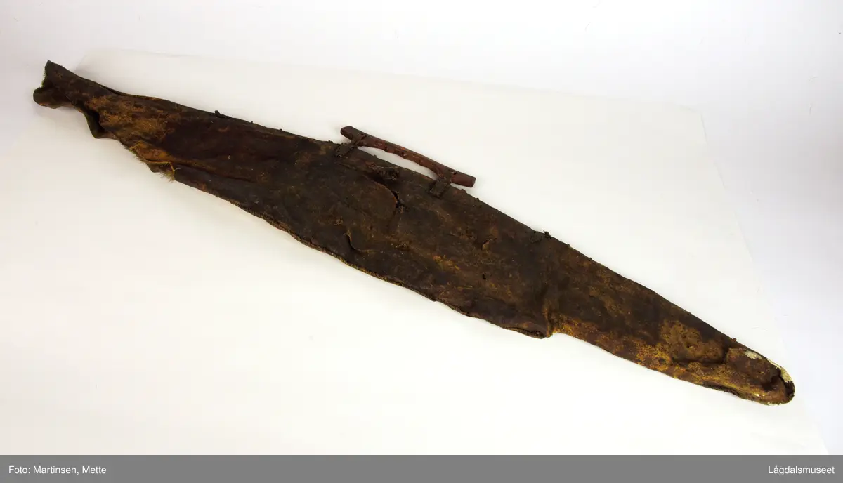 Bueveske av skinn. Pelssiden inn, sydd med sene Plass for piler og bue. Bærehåndtak av tre festet til buevesken med skinnsnorer, sydd med sene. I nederkant ca. 32 cm lang sprekk til åpning.