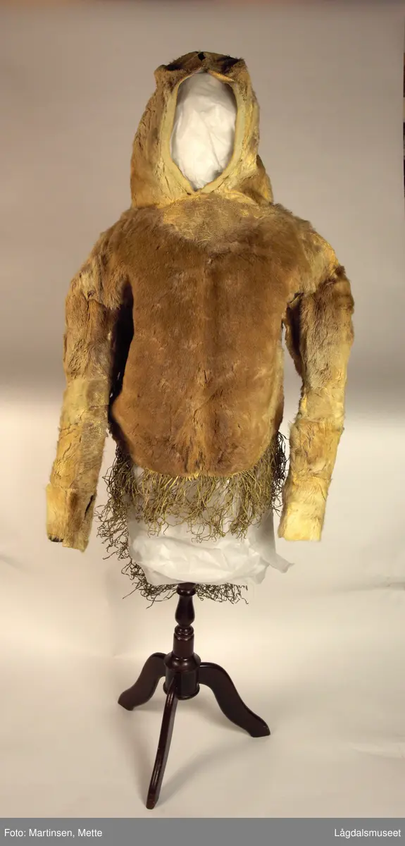 Anorakk eller parkas sydd av reinskinn. Pelssiden ut. Påsydd hette. Bakstykket lenger enn fronten. Langs nederkant påsydde frynser av skinn.