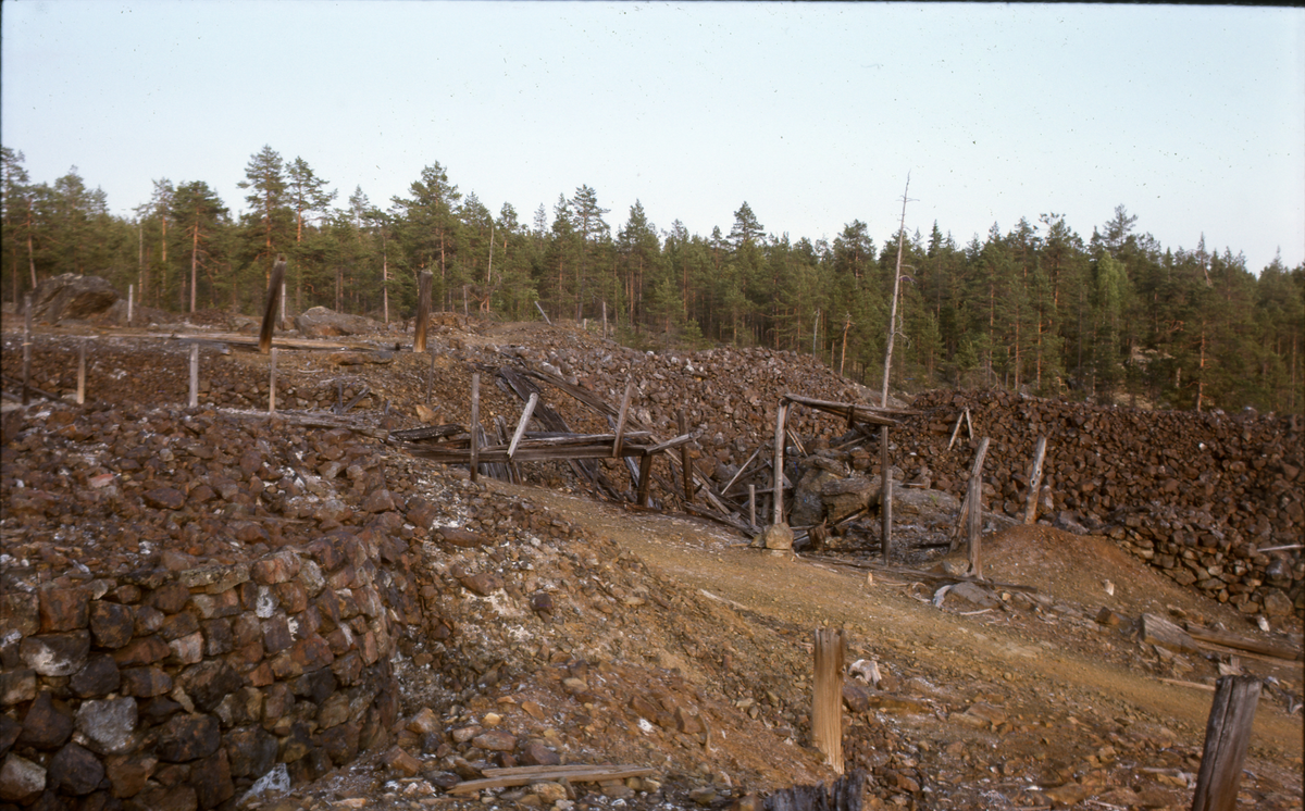 Flätsbo gruva. Bruten mark och en grusväg, tallskog i bakgrunden.