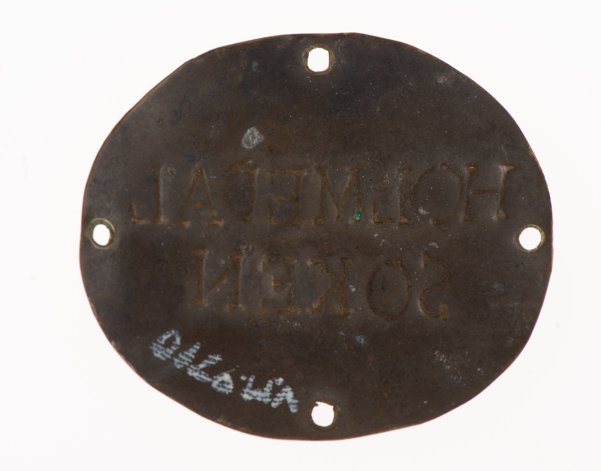 Oval mässingsbricka med fyra hål för att fästa i kläderna med sytråd. Holmedal socken.