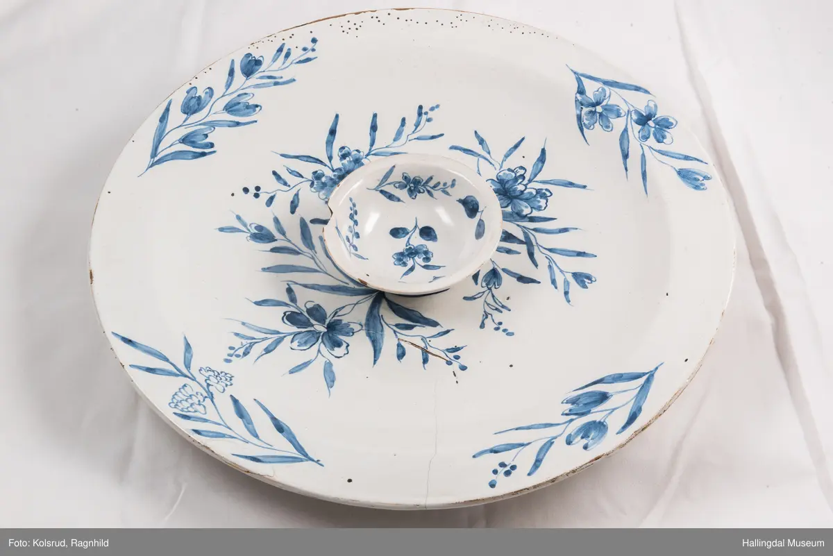 Fajanse, melkehvitt, rundt. Undersiden vid, markert fotring. Svakt skrånende side, smal rand. Dekorasjoner i blått, omkring smørgruben 3 grener med smale blad og hver en blomst. Langs randen 4 grener med forskjellige blomster. R.B.S.