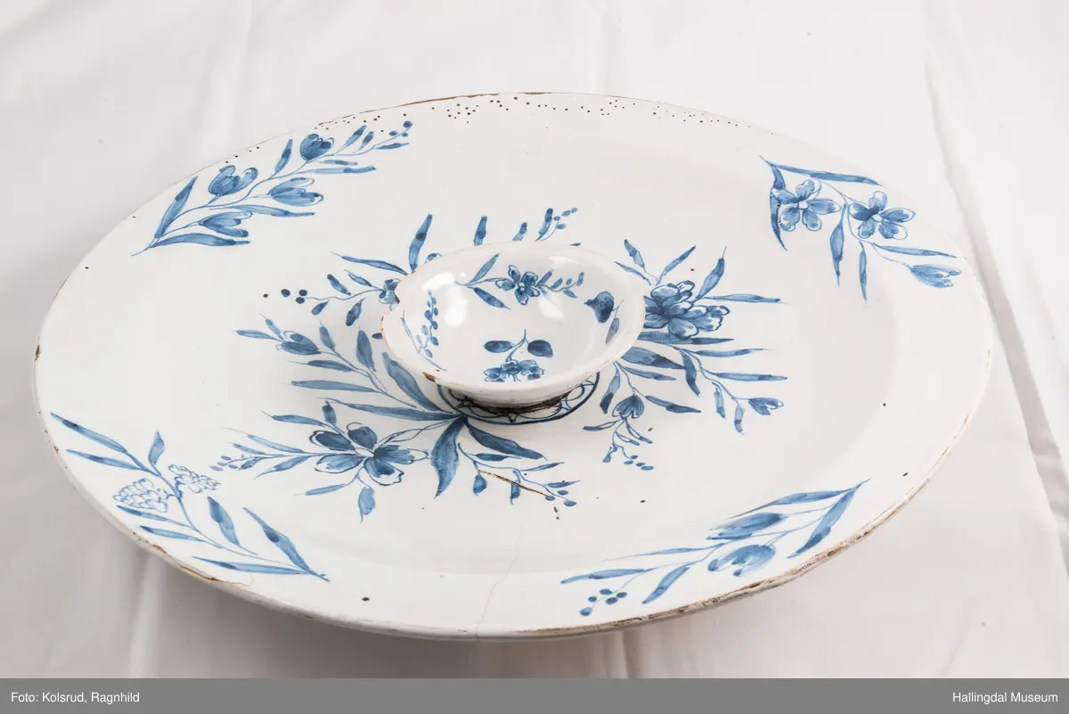 Fajanse, melkehvitt, rundt. Undersiden vid, markert fotring. Svakt skrånende side, smal rand. Dekorasjoner i blått, omkring smørgruben 3 grener med smale blad og hver en blomst. Langs randen 4 grener med forskjellige blomster. R.B.S.