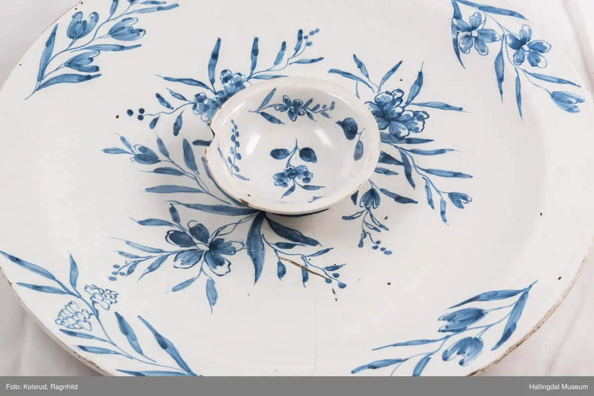 Fajanse, melkehvitt, rundt. Undersiden vid, markert fotring. Svakt skrånende side, smal rand. Dekorasjoner i blått, omkring smørgruben 3 grener med smale blad og hver en blomst. Langs randen 4 grener med forskjellige blomster. R.B.S.