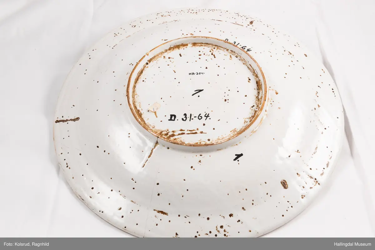 Fajanse, melkehvitt, rundt. Undersiden vid, markert fotring. Svakt skrånende side, smal rand. Dekorasjoner i blått, omkring smørgruben 3 grener med smale blad og hver en blomst. Langs randen 4 grener med forskjellige blomster. R.B.S.