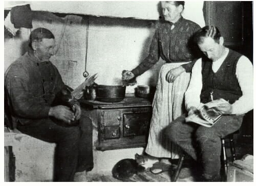 Familjebild. 1923 ca. Bengt-Karl och Karolina med sonen August vid spisen i köket, Hulegård, Krogsered. Karolina rör i grytan, Bengt-Karl sitter med en katt i knät och en vid fötterna. August bläddrar i en tidning.
Bilden tagen med magnesiumblixt.