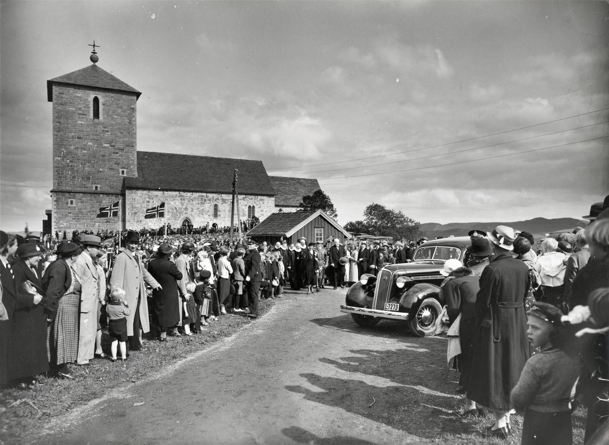 Kronprins Olav og kronprinsesse Martha på vei bort fra Olavskirka på Avaldsnes, fulgt av prester og en stor folkemengde. Bilen er en 1936-modell Studebaker med registreringsnummer L-7270.