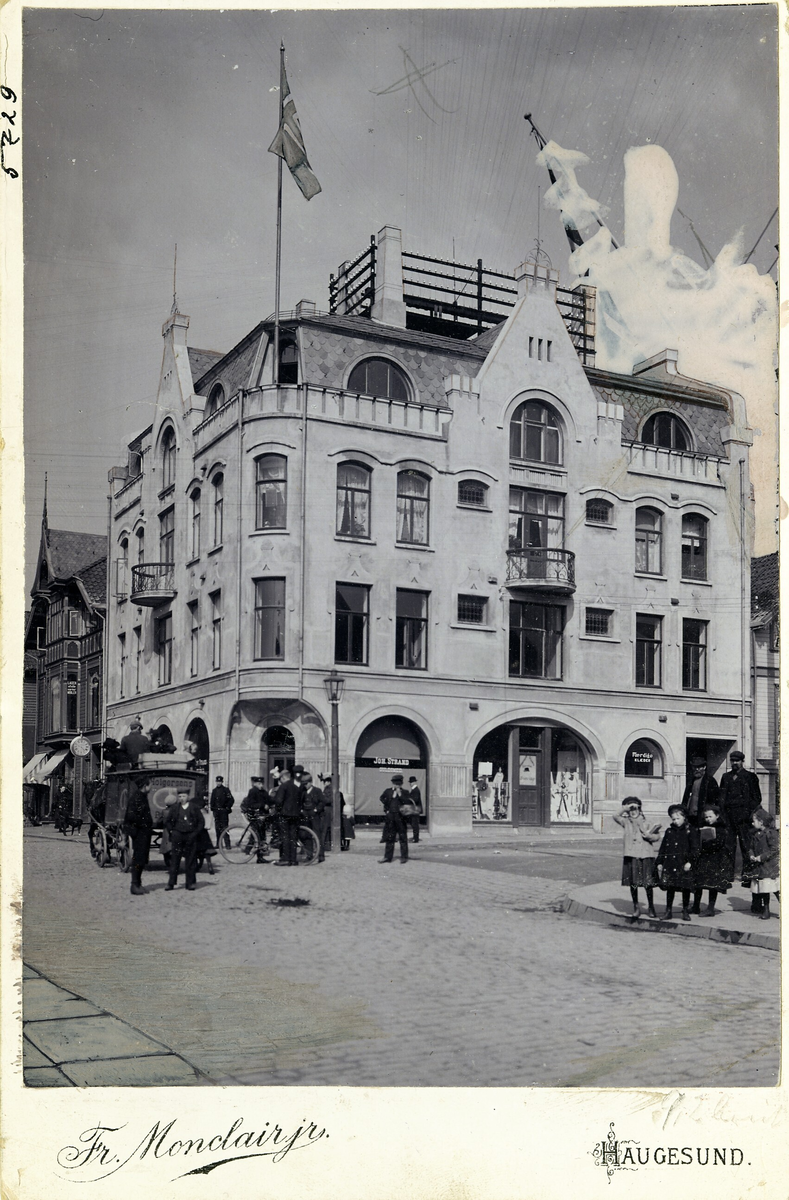 Postkort og retusjert kabinettkort av Telefonsentralen i Haraldsgata 161, Haugesund. I 1. etg. holdt en klesforretning og gullsmeden Joh. Strand til. Hestevogna til Holgersens bakeri passerer og flere titter på fotografen.