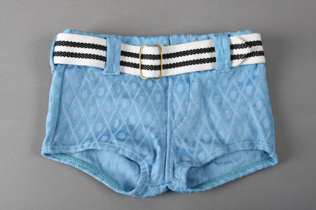 Badebukse til gutt. Laget av 100% nylon. Lys blå tekstil med innvevd ruterdekor med oval form inni. Badebuksen har påsydde beltehemper og et hvitt belte med svarte striper. Beltespenne i forgylt metall. Nylonforing inni. Det er påsydd en liten lomme inni. Størrelse 14.