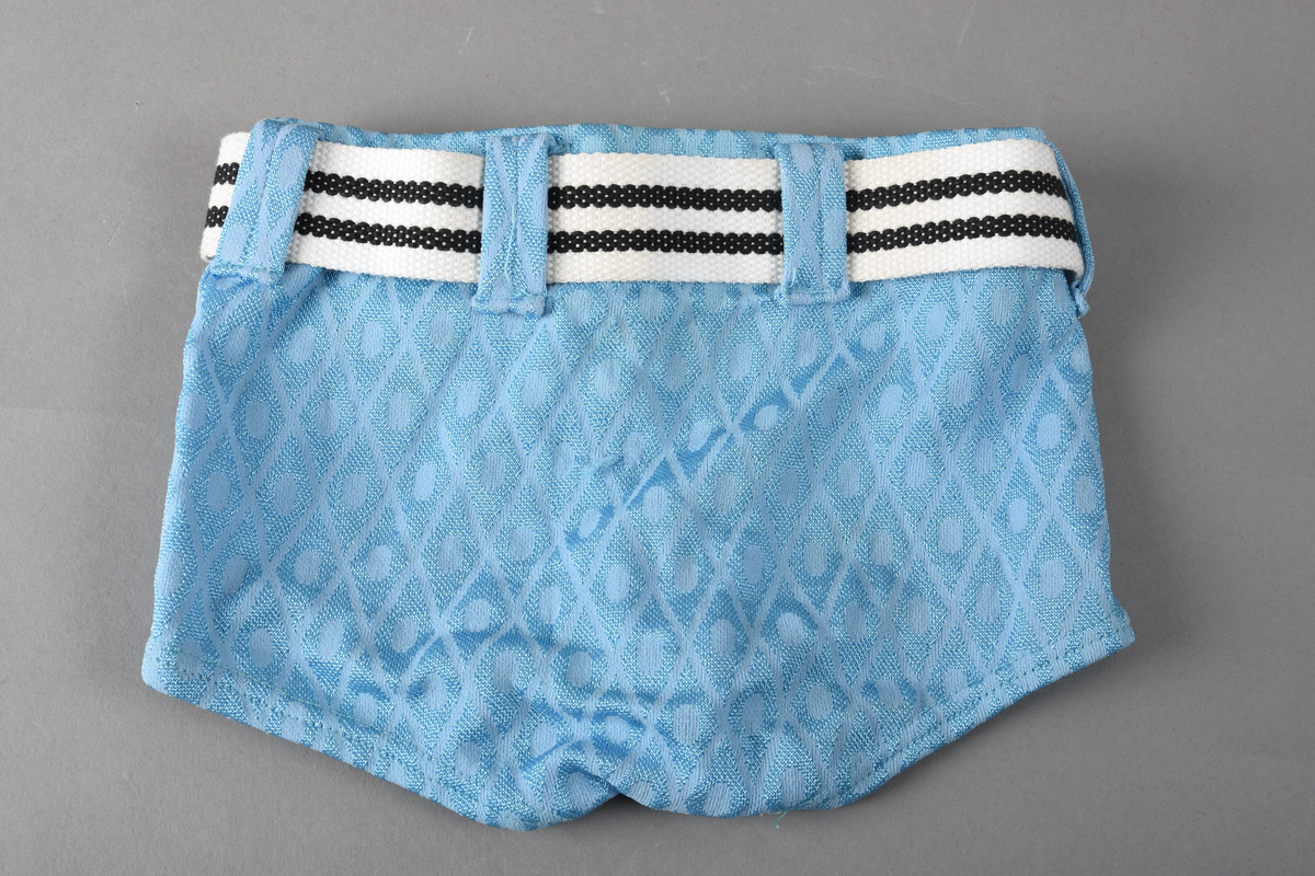 Badebukse til gutt. Laget av 100% nylon. Lys blå tekstil med innvevd ruterdekor med oval form inni. Badebuksen har påsydde beltehemper og et hvitt belte med svarte striper. Beltespenne i forgylt metall. Nylonforing inni. Det er påsydd en liten lomme inni. Størrelse 14.