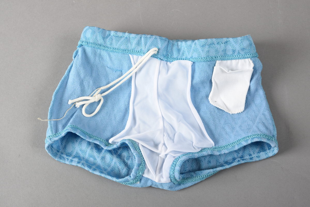 Badebukse til gutt. Laget av 100% nylon. Lys blå tekstil med innvevd ruterdekor med oval form inni. Badebuksen har påsydde beltehemper og et hvitt belte med svarte striper. Beltespenne i forgylt metall. Nylonforing inni. Det er påsydd en liten lomme inni. Størrelse 14.