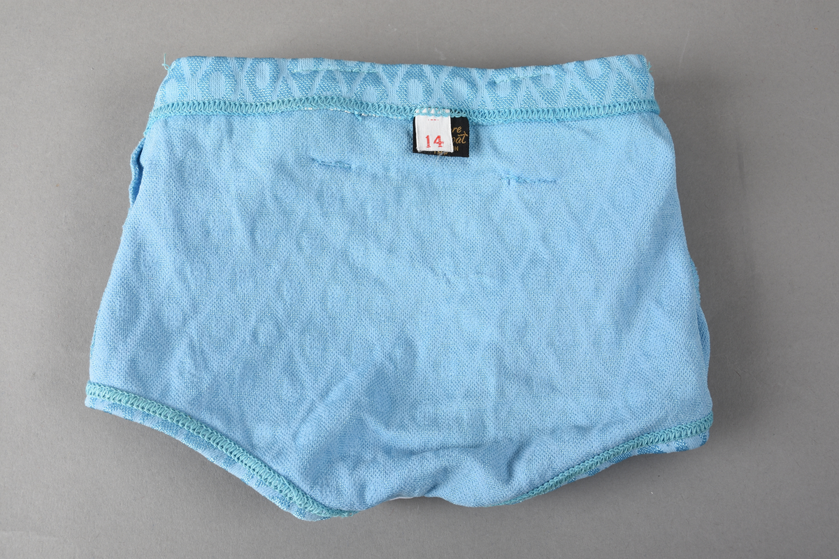 Badebukse til gutt. Laget av 100% nylon. Lys blå tekstil med innvevd ruterdekor med oval form inni. Badebuksen har påsydde beltehemper og et hvitt belte med svarte striper. Beltespenne i forgylt metall. Nylonforing inni. Det er påsydd en liten lomme inni. Størrelse 14.
