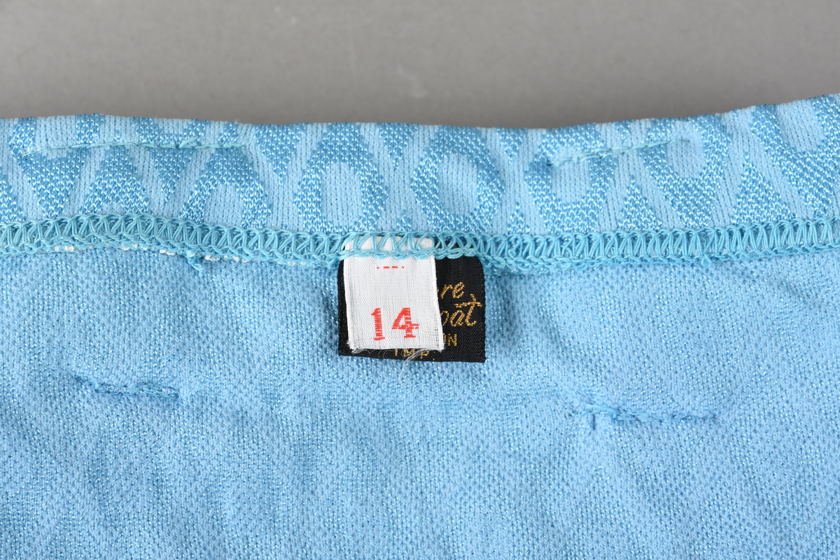 Badebukse til gutt. Laget av 100% nylon. Lys blå tekstil med innvevd ruterdekor med oval form inni. Badebuksen har påsydde beltehemper og et hvitt belte med svarte striper. Beltespenne i forgylt metall. Nylonforing inni. Det er påsydd en liten lomme inni. Størrelse 14.