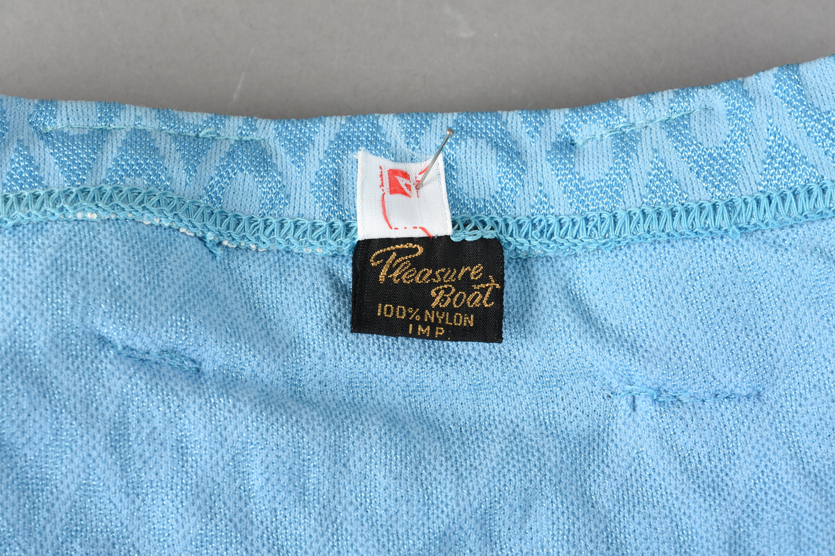 Badebukse til gutt. Laget av 100% nylon. Lys blå tekstil med innvevd ruterdekor med oval form inni. Badebuksen har påsydde beltehemper og et hvitt belte med svarte striper. Beltespenne i forgylt metall. Nylonforing inni. Det er påsydd en liten lomme inni. Størrelse 14.