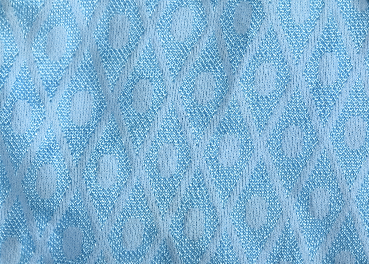 Badebukse til gutt. Laget av 100% nylon. Lys blå tekstil med innvevd ruterdekor med oval form inni. Badebuksen har påsydde beltehemper og et hvitt belte med svarte striper. Beltespenne i forgylt metall. Nylonforing inni. Det er påsydd en liten lomme inni. Størrelse 14.