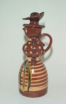 Kanna med hänkel och propp i lergods formad som en kvinnokropp med hornmålat mönster samt kjol med förkläde.