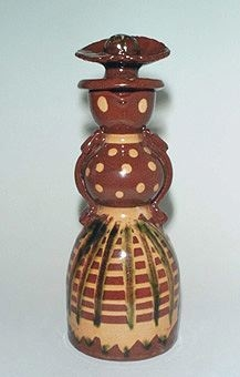 Kanna med hänkel och propp i lergods formad som en kvinnokropp med hornmålat mönster samt kjol med förkläde.