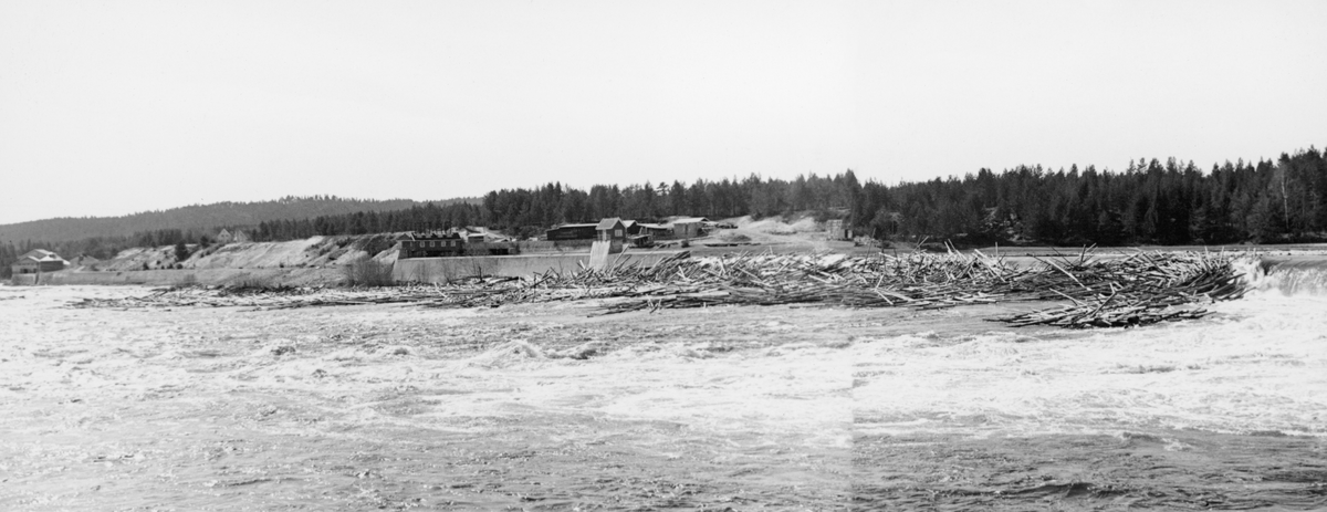 Kraftverksdammen ved Skjefstadfossen i Heradsbygda i Elverum.  Fotografiet er tatt i slutten av april 1942, etter at vårflommen og tømmerfløtinga hadde begynt å sette sitt preg på Glommavassdraget.  Bildet er tatt fra elvas østre bredd mot elveleiet like nedenfor dammen, der det hadde lagt seg tømmervaser på steinører som kun var dekt av et grunt vannlag.  I bakgrunnen til høyre ser vi inntaksdammen med tilhørende bygninger.  Derfra gikk turbinrørene til kraftverket, som vi ser til venstre i dette bildet (bortimot 500 meter lengre nede ved elvas vestbredd).  Elveskråningene nedenfor dammen preges av vegetasjonsfattige, bratte sandflater.  I Glomma fellesfløtingsforenings album nr. 13 er dette fotografiet montert tett inntil et annet, unummerert bilde, som en slags vidvinkelmontasje.  Vinteren før dette fotografiet ble tatt foregikk det anleggsarbeid på dammen (jfr. SJF. 1989-03947-49). 