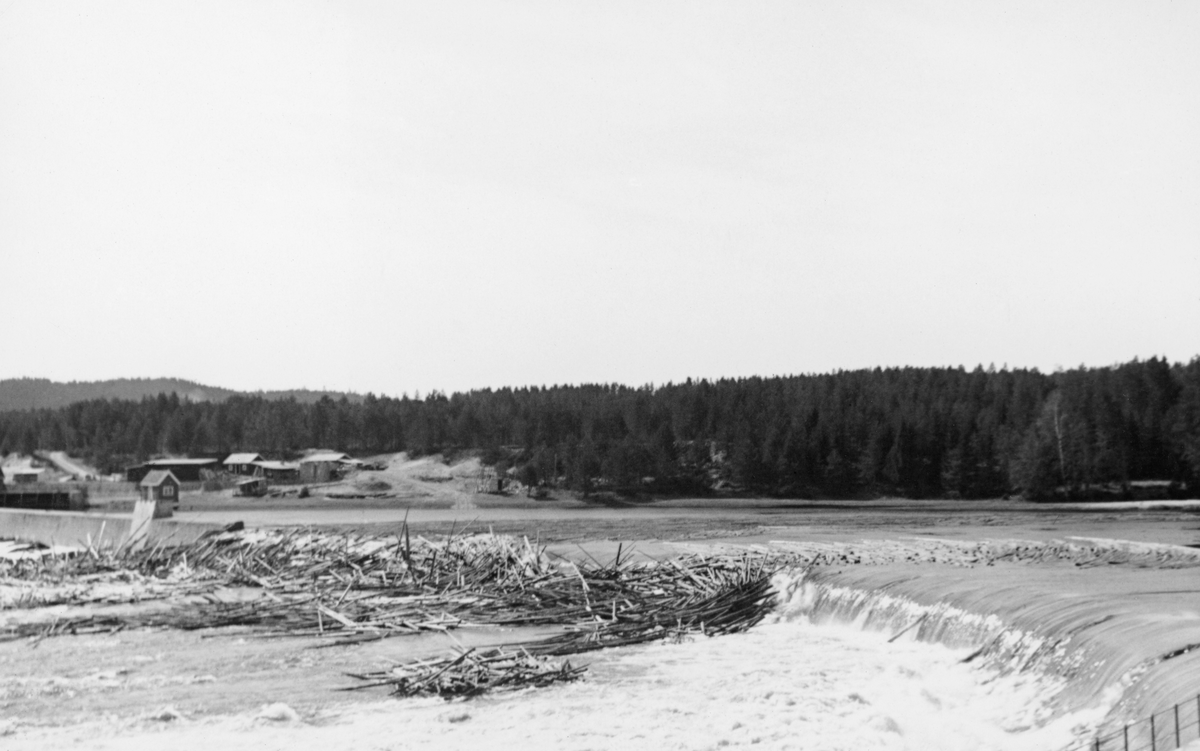 Kraftverksdammen ved Skjefstadfossen i Heradsbygda i Elverum.  Fotografiet er tatt i slutten av april 1942, etter at vårflommen og tømmerfløtinga hadde begynt å sette sitt preg på Glommavassdraget.  Bildet er tatt fra et oppmurt utsiktspunkt på elvas østre bredd mot dammen elveleiet like nedenfor.  I forgrunnen skimter vi noe av rekkverket på dette utsiktspunktet.  I forgrunnen, like nedenfor, ser vi det kvitskummende vannet som nettopp har passert damkrona i den djupeste og mest strømrike delen av elveleiet.  Midt i elveleiet hadde det lagt seg tømmervaser på steinører som kun var dekt av et grunt vannskikt.  Vasedannelsen fortsetter også ovenfor damkrona.  I bakgrunnen til venstre skimter vi noe av inntaksdammen, der turbinrørene til kraftverket, som lå litt lengre nede ved elvas vestbredd.  På og bakenfor inntakstdammen lå en del mindre bygninger.  Landskapet i bakgrunnen preges av barskog. Vinteren før dette fotografiet ble tatt foregikk det anleggsarbeid på dammen (jfr. SJF. 1989-03947-49). 