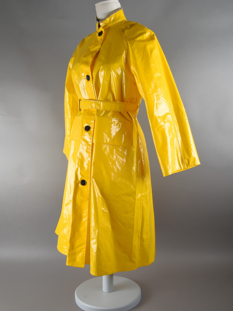 Regnfrakk av plast-stoff. Trenchcoat-aktig. Fire svarte knapper med knapphull foran. To lommer med klaff over på framsiden, på hver side. Belte med svart beltespenne som henger i to beltehemper på hver side, bak. Lange ermer. Splitt på baksiden, nede. Innvendig krave av kordfløyel. Hempe til oppheng i nakken. Det er knytt fast en plasttråd i hempen. Merkelapp med tekst på festet under hempen.