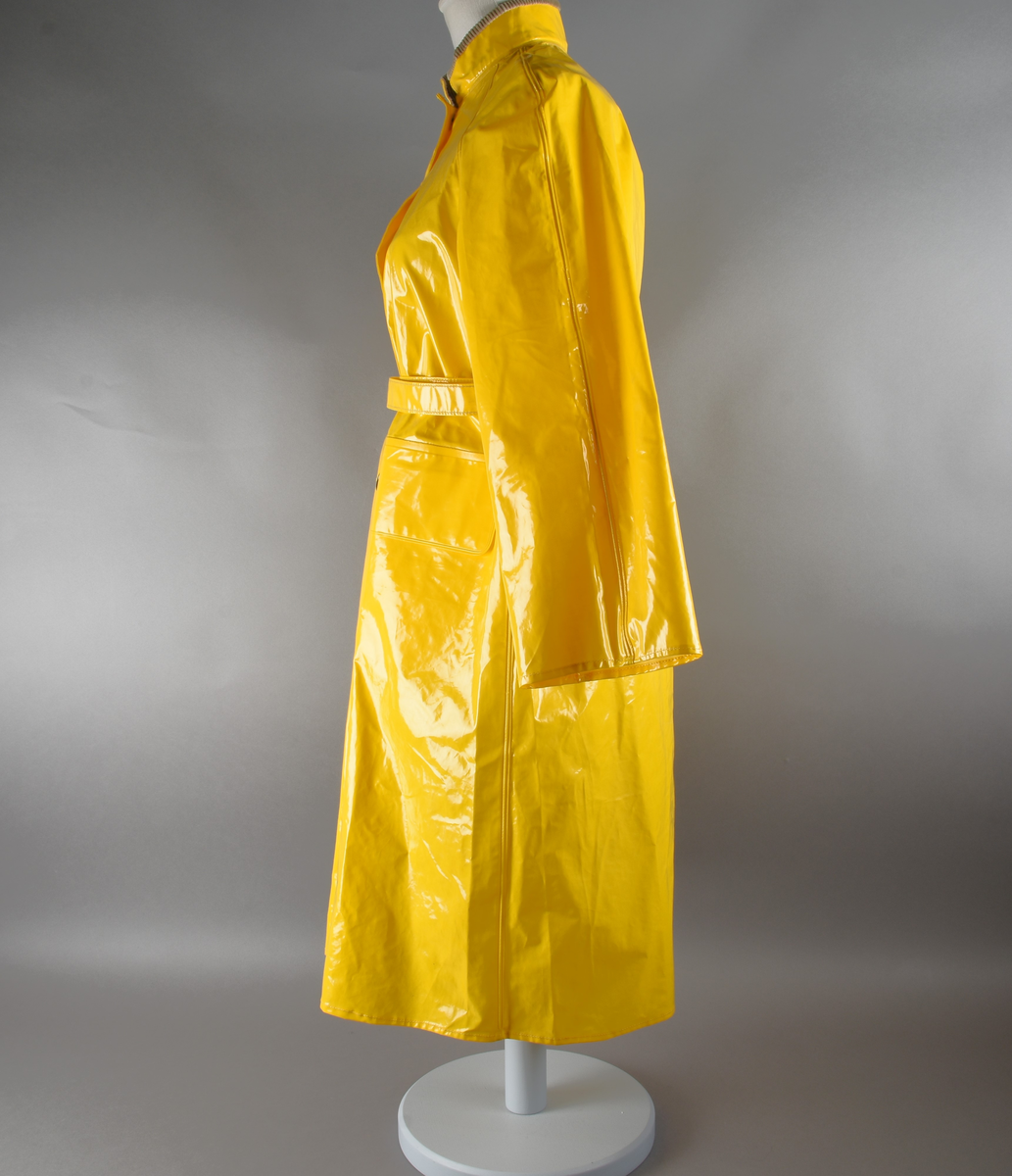 Regnfrakk av plast-stoff. Trenchcoat-aktig. Fire svarte knapper med knapphull foran. To lommer med klaff over på framsiden, på hver side. Belte med svart beltespenne som henger i to beltehemper på hver side, bak. Lange ermer. Splitt på baksiden, nede. Innvendig krave av kordfløyel. Hempe til oppheng i nakken. Det er knytt fast en plasttråd i hempen. Merkelapp med tekst på festet under hempen.
