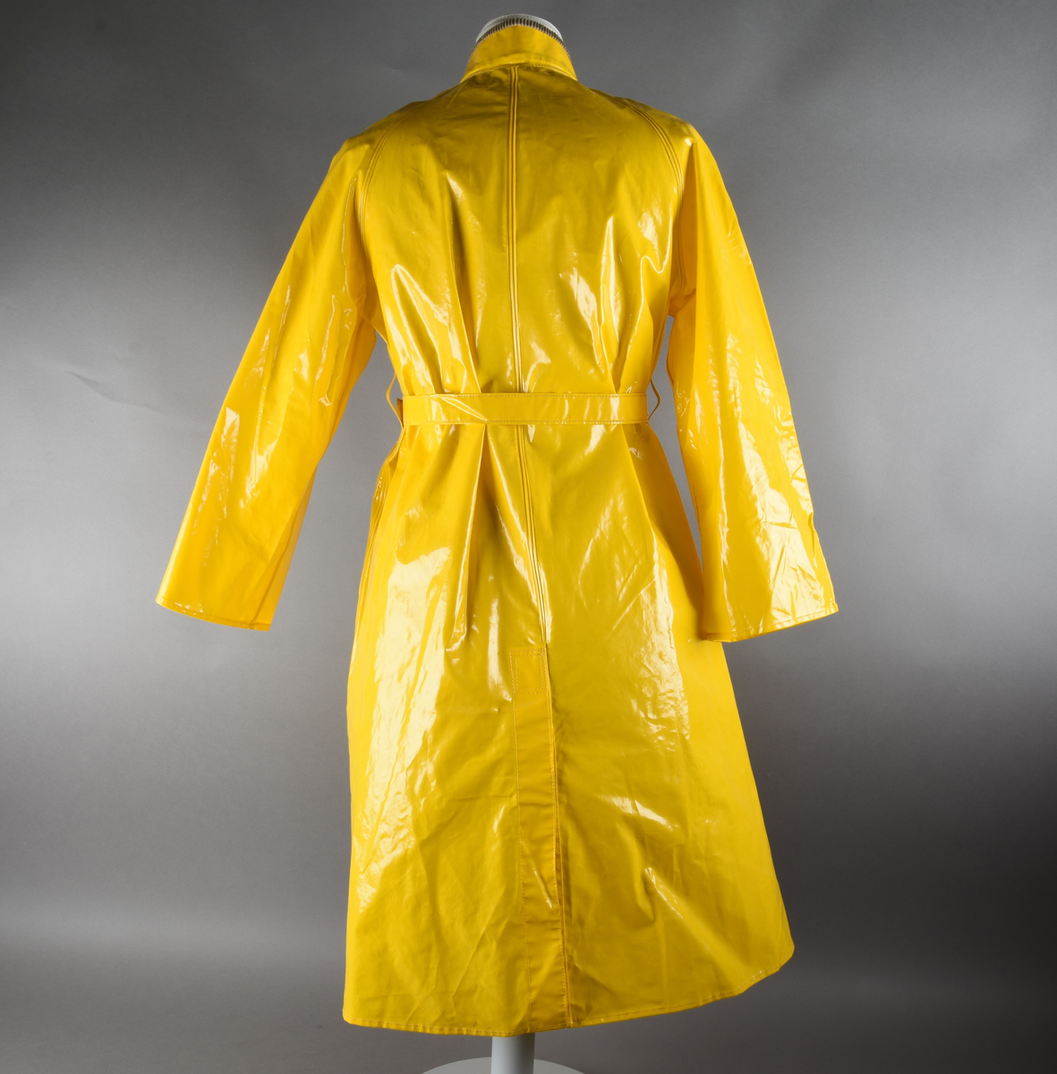 Regnfrakk av plast-stoff. Trenchcoat-aktig. Fire svarte knapper med knapphull foran. To lommer med klaff over på framsiden, på hver side. Belte med svart beltespenne som henger i to beltehemper på hver side, bak. Lange ermer. Splitt på baksiden, nede. Innvendig krave av kordfløyel. Hempe til oppheng i nakken. Det er knytt fast en plasttråd i hempen. Merkelapp med tekst på festet under hempen.