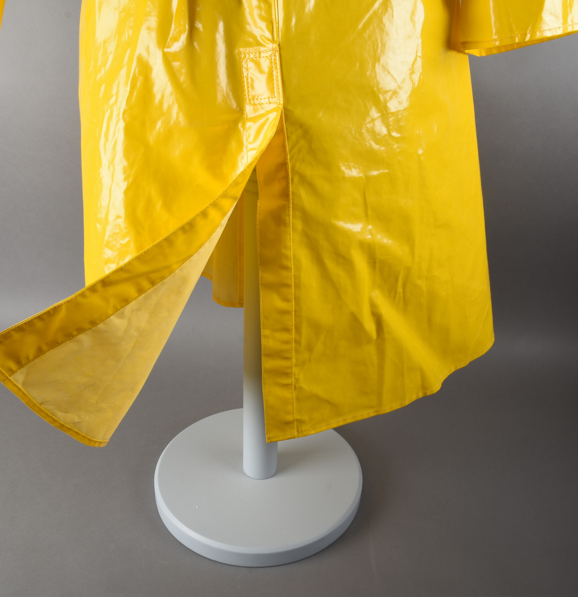 Regnfrakk av plast-stoff. Trenchcoat-aktig. Fire svarte knapper med knapphull foran. To lommer med klaff over på framsiden, på hver side. Belte med svart beltespenne som henger i to beltehemper på hver side, bak. Lange ermer. Splitt på baksiden, nede. Innvendig krave av kordfløyel. Hempe til oppheng i nakken. Det er knytt fast en plasttråd i hempen. Merkelapp med tekst på festet under hempen.