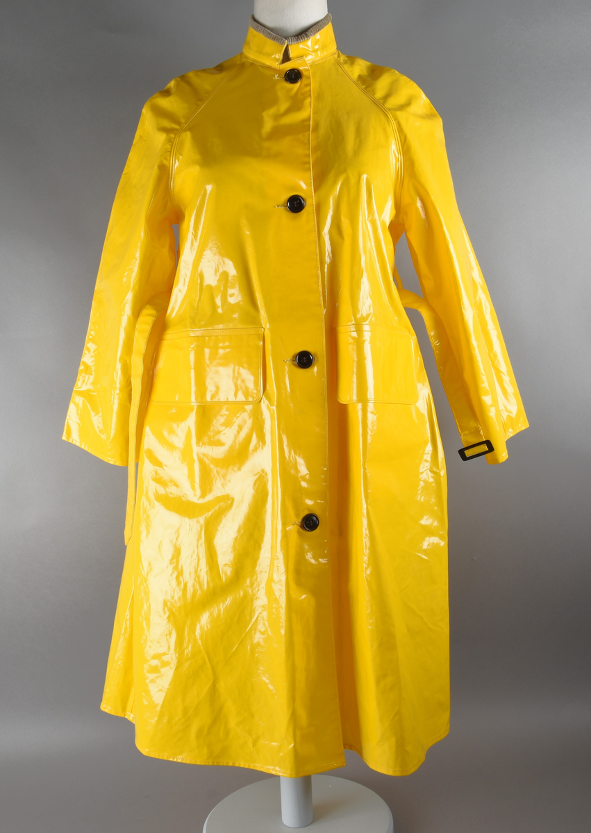 Regnfrakk av plast-stoff. Trenchcoat-aktig. Fire svarte knapper med knapphull foran. To lommer med klaff over på framsiden, på hver side. Belte med svart beltespenne som henger i to beltehemper på hver side, bak. Lange ermer. Splitt på baksiden, nede. Innvendig krave av kordfløyel. Hempe til oppheng i nakken. Det er knytt fast en plasttråd i hempen. Merkelapp med tekst på festet under hempen.