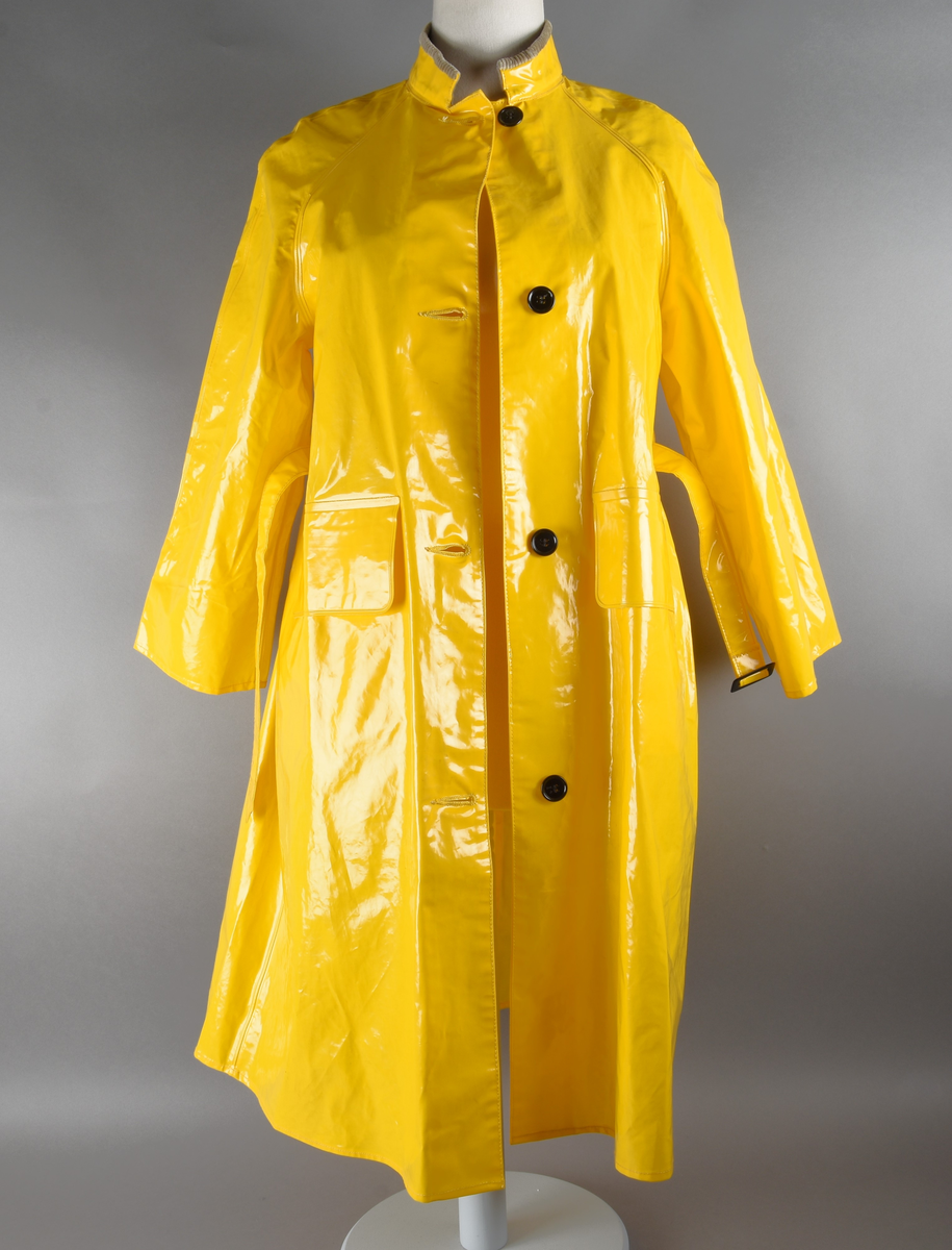 Regnfrakk av plast-stoff. Trenchcoat-aktig. Fire svarte knapper med knapphull foran. To lommer med klaff over på framsiden, på hver side. Belte med svart beltespenne som henger i to beltehemper på hver side, bak. Lange ermer. Splitt på baksiden, nede. Innvendig krave av kordfløyel. Hempe til oppheng i nakken. Det er knytt fast en plasttråd i hempen. Merkelapp med tekst på festet under hempen.