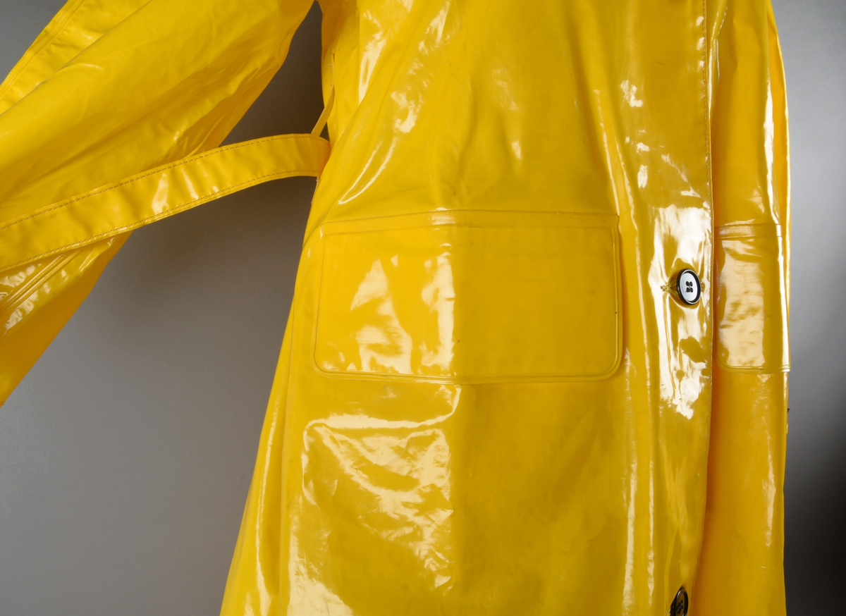 Regnfrakk av plast-stoff. Trenchcoat-aktig. Fire svarte knapper med knapphull foran. To lommer med klaff over på framsiden, på hver side. Belte med svart beltespenne som henger i to beltehemper på hver side, bak. Lange ermer. Splitt på baksiden, nede. Innvendig krave av kordfløyel. Hempe til oppheng i nakken. Det er knytt fast en plasttråd i hempen. Merkelapp med tekst på festet under hempen.