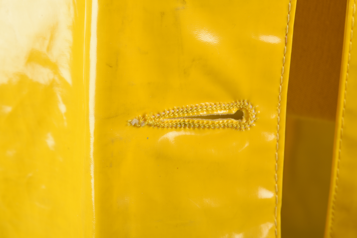 Regnfrakk av plast-stoff. Trenchcoat-aktig. Fire svarte knapper med knapphull foran. To lommer med klaff over på framsiden, på hver side. Belte med svart beltespenne som henger i to beltehemper på hver side, bak. Lange ermer. Splitt på baksiden, nede. Innvendig krave av kordfløyel. Hempe til oppheng i nakken. Det er knytt fast en plasttråd i hempen. Merkelapp med tekst på festet under hempen.