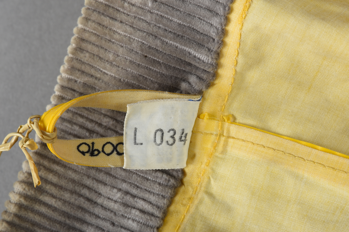 Regnfrakk av plast-stoff. Trenchcoat-aktig. Fire svarte knapper med knapphull foran. To lommer med klaff over på framsiden, på hver side. Belte med svart beltespenne som henger i to beltehemper på hver side, bak. Lange ermer. Splitt på baksiden, nede. Innvendig krave av kordfløyel. Hempe til oppheng i nakken. Det er knytt fast en plasttråd i hempen. Merkelapp med tekst på festet under hempen.