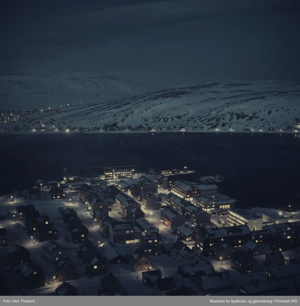 Det ligger snø i Hammerfest og og på fjellene rundt. Det er lys i husene og gatebelysningen er på. Utsikten strekker seg fra Salenfjellet, langs Fuglenesveien og  helt bort til Storfjellet.