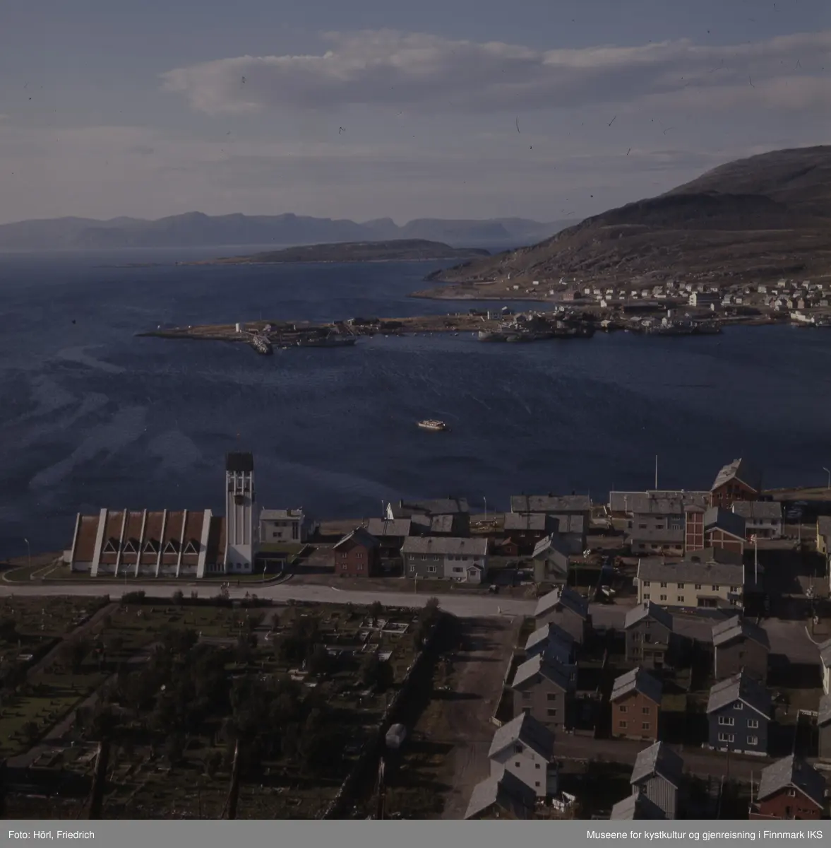 Det er sommer i Hammerfest. Fra Salenfjellet ser man ned til kirkegården, den gjenreiste protestantiske kirka og brannstasjonen på Hauen. En båt ligger i innseilinga til havna. I øvre delen av bildet ser man bydelen Fuglenes og Rossmollbukta med gjenreisningsbebyggelsen og slipp. Ute i Sørøysundet kan man se ut over Melkøya og Sørøya i bakgrunn.