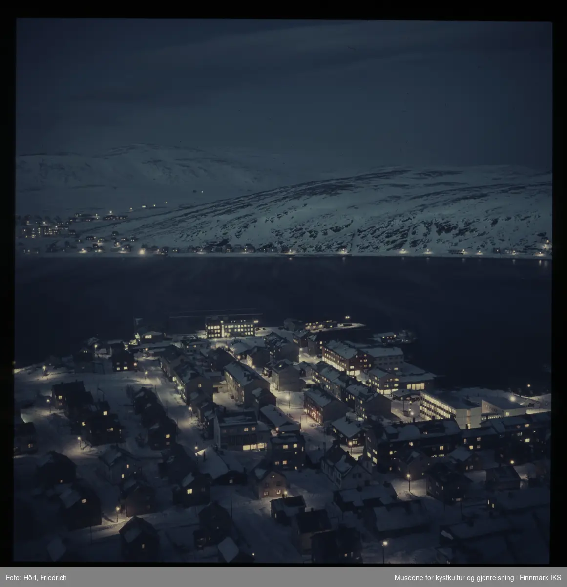 Det ligger snø i Hammerfest og og på fjellene rundt. Det er lys i husene og gatebelysningen er på. Utsikten strekker seg fra Salenfjellet, langs Fuglenesveien og  helt bort til Storfjellet.