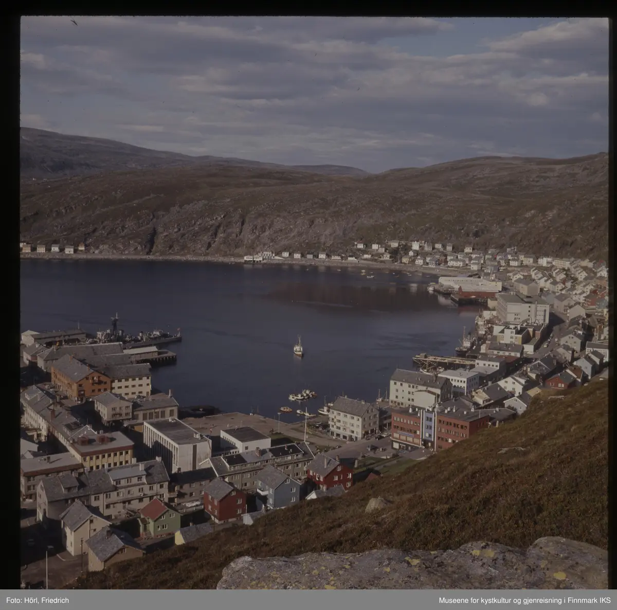 Det er sommer i Hammerfest og fra Salenfjellet ser man utover byens sentrum med rådhus, bankbygg og Findusfabrikken helt bort til Hauans materialandel ved munningen til Storelva og bydelen Molla bak den. Byen er gjenreist og fargerike hus med skifertak preger bybildet. I havna liger det ulike typer båter. Marinefartøyet på bildet er en norsk fregatt, mest sannsynlig KNM Tromsø eller KNM Haugesund.