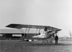 Militärt flygplan Lohner B.III märkt nr 14.08 tillhörande Österrike-Ungern står på ett flygfält. En man sitter i flygplanet. Vid flygplansnosen står en man. Flyghangar i bakgrunden. Vy framifrån.