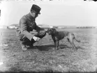 En militär från Österrike-Ungern sitter på huk bredvid en hund på ett flygfält. Byggnader i bakgrunden. Vy från sidan.