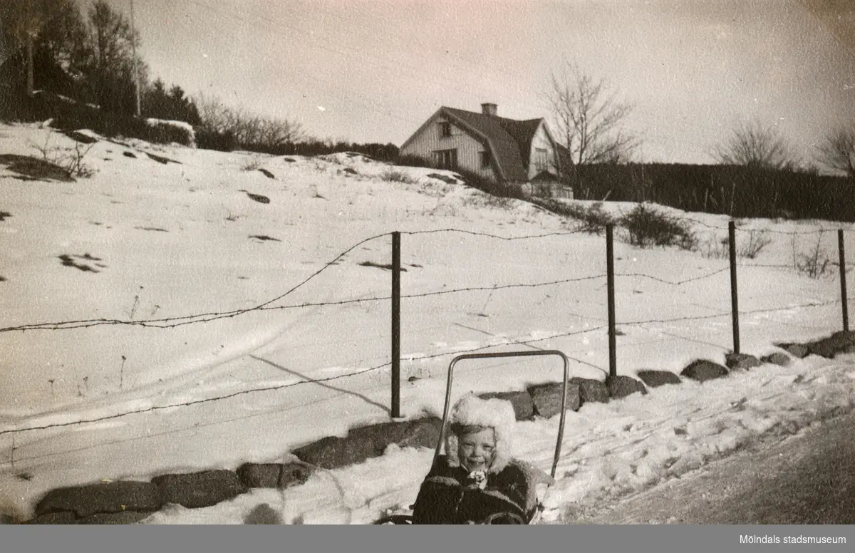 Eva Pettersson (född 1944, gift Kempe) sitter i kärran i det snötäckta Sagered, februari 1946. Familjen är på väg till mormor som bor i Stretered.