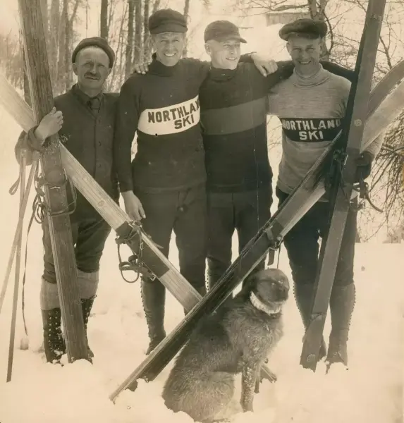 Fire skihoppere i vinterklær. luer og kraftige treski, poserer sammen med hund