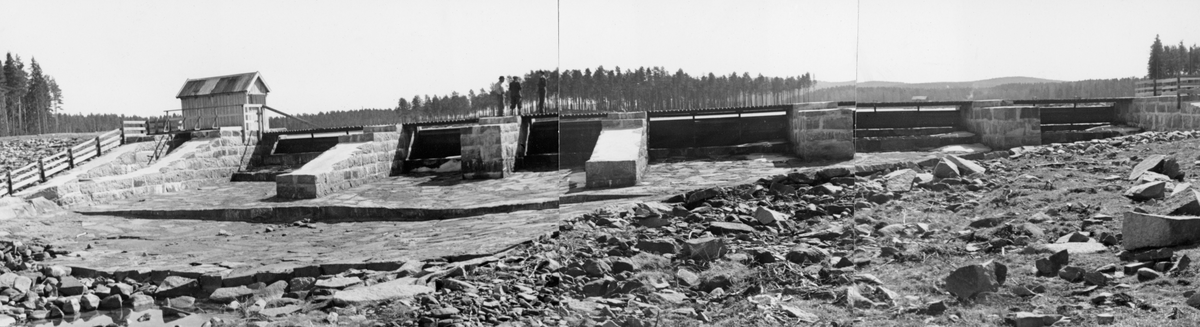 Fra Rokosjødammen i Løten i Hedmark, fotografert nedenfra, altså i motstrøms retning. Dammen ligger ved Rokoelvas (Svartelvas) utløp fra den vestre enden av sjøen. Her var den 4 kilometer lange og opptil 1,5 kilometer brede Rokosjøen regulert ved hjelp av en dam med kar som var murt av kvadret naturstein. På den tredelte panoramamontasjen dette fotografiet er den midtre delen av ser vi seks løp (jfr. SJF.1989-03761 og SJF.1989-03763). I tillegg var det antakelig et avløp under det lille bordkledde bindingsverkshuset på damkrona, til venstre i bildet, som antas å ha vært et lukehus. På tvers av løpene gikk det ei trebru. Fotografiet ble tatt 1. mai 1942. På dette tidspunktet var sjøen nedtappet, slik at det ikke rant vann gjennom dammen.

Rokosjøen ble ifølge Gunnar Sætrens bok «Beskrivelse af Glommen» (1904) regulert med en dam allerede i 1830, Målet var å husholdere med vannet til beste for kverner, møller, sagbruk og etter hvert også andre industriforetak ved Svartelva, som hadde mange dammer og fall før den etter snaut to mil nådde Åkersvika i Mjøsa. Den Rokosjødammen vi ser på dette fotografiet ble bygd i 1937-38 - jfr. fanen «Opplysninger».

I Glomma fellesfløtingsforenings album inngår dette motivet i en todelt montasje - jfr. SJF.1989-03762.